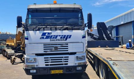 A white Volvo semi-truck with “Scrap Salvage” written in blue on its cab is parked in a sunny industrial yard, facing forward. A white Volvo semi-truck with "Scrap Salvage" written in blue on its cab is parked in a sunny industrial yard, facing forward.